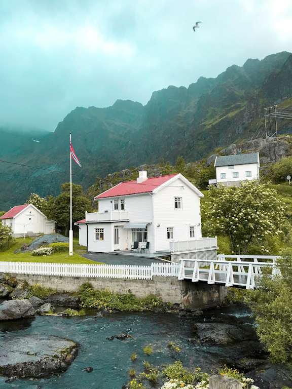 A white house with a red roof is situated by a body of water with a bridge leading to it.