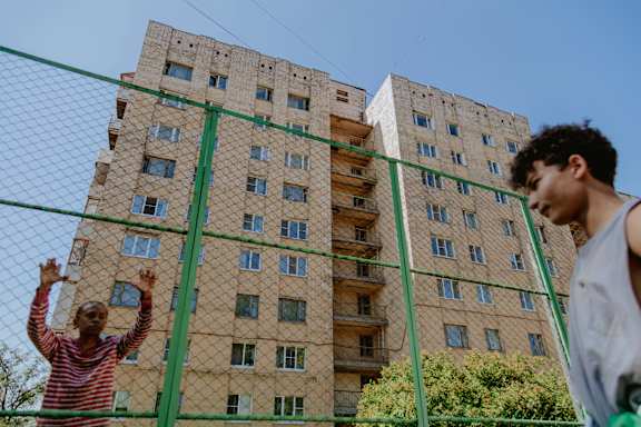 Two people standing in front of a tall building with a green fence.