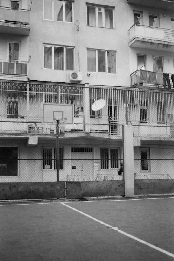 A black and white photo of a basketball court in front of a white building with balconies.