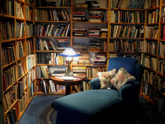 A cozy reading nook with a blue chair, a table with a lamp, and bookshelves filled with books.