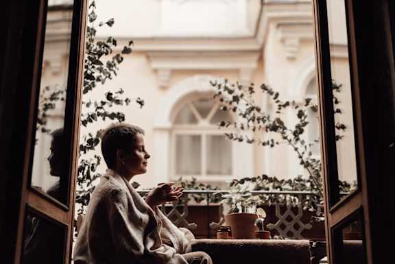 A person is sitting by a window with a view of a building and plants.