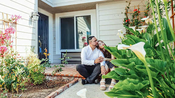 A couple is sitting on the ground outside a house, surrounded by plants and flowers.