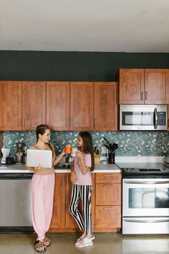Two women in a kitchen, one holding a laptop and the other holding a mug.