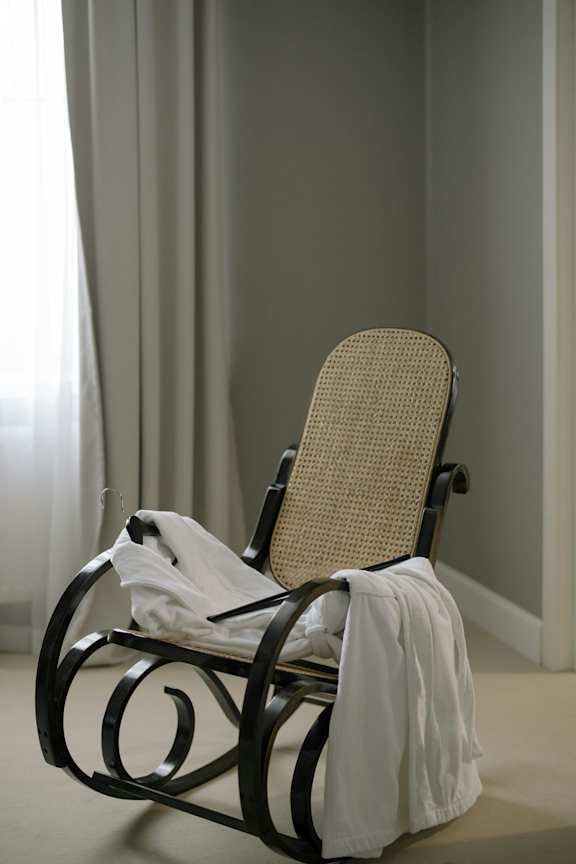 A chair with a woven seat and a white cloth draped over the back is in front of a window with sheer curtains.