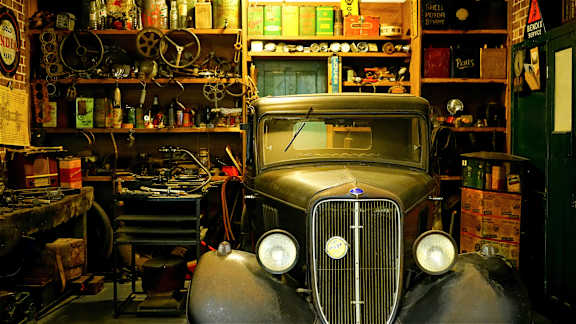 An old car is parked in a garage with a lot of tools and equipment.