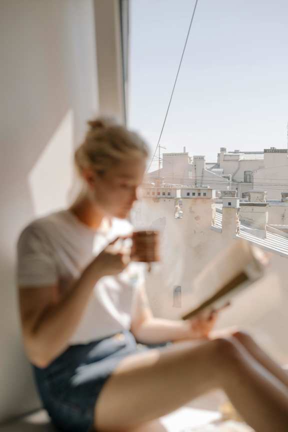 A woman is sitting by a window reading a book.