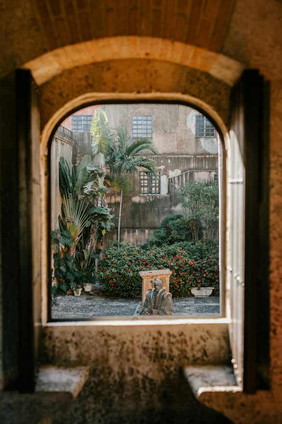 A view through an arched window into a courtyard with plants and a fountain.