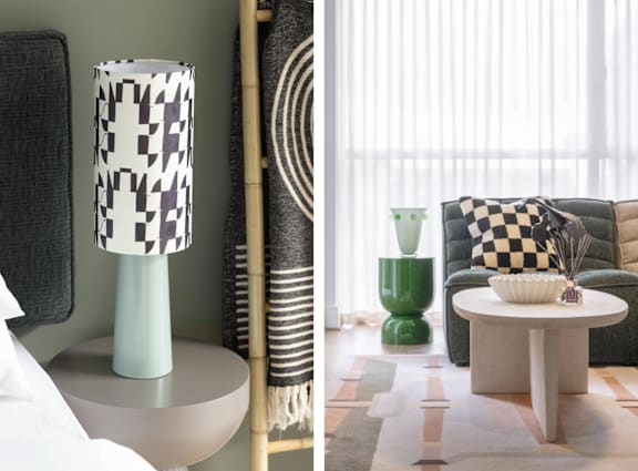 A lamp with a black and white checkered shade sits on a table next to a green vase and a checkered pillow.
