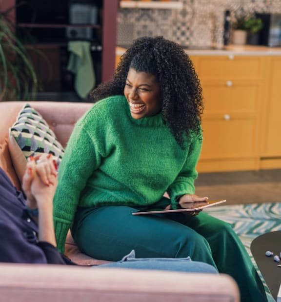A woman with curly hair is sitting on a couch, wearing a green sweater and holding a tablet.
