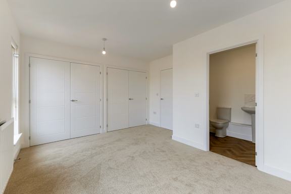 A room with white walls and a carpeted floor with a toilet visible in the adjoining room.