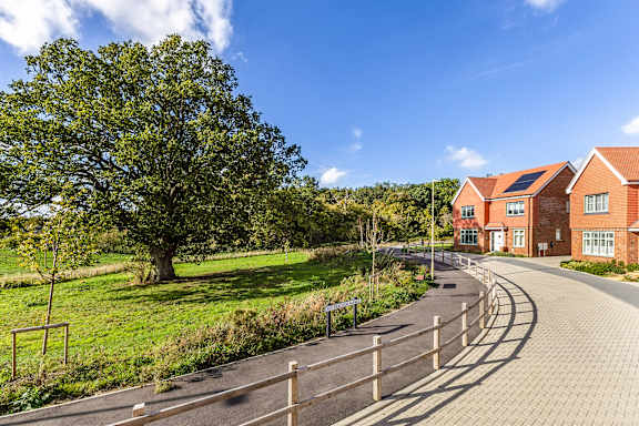 A curved pathway with a wooden fence and a green tree on the left, leading to a building with red brick walls and white windows.
