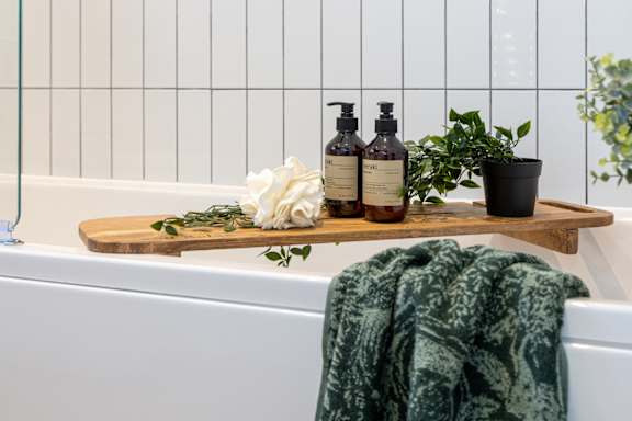 A bathroom with a white tub, a wooden tray with two bottles, a potted plant, and a green towel.