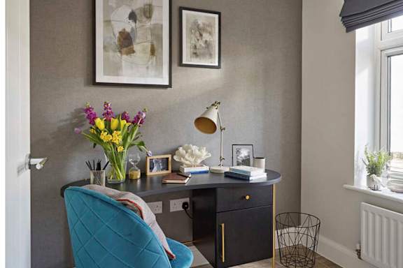 A blue chair sits in front of a desk with a lamp and a vase of flowers.