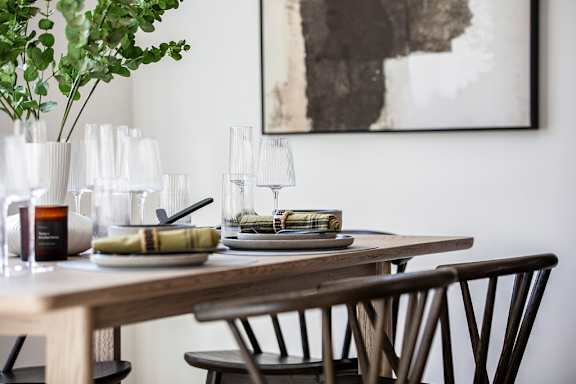 A dining table set with plates and glasses with a potted plant and a painting in the background.