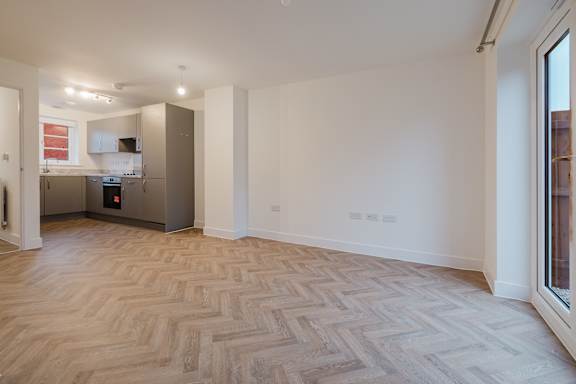 A spacious room with a herringbone parquet floor and a kitchen in the background.