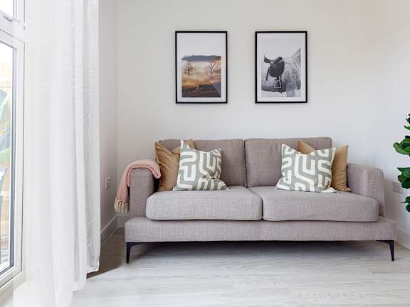 A grey couch with two pillows and a pink blanket sits in a room with two framed pictures on the wall.