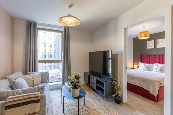 A living room with a couch, a chair, a coffee table, a television, and a bedroom with a bed and a nightstand.