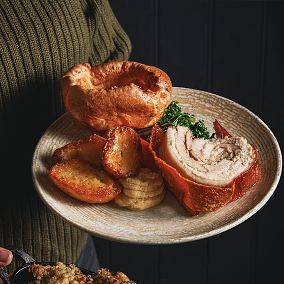 A person is holding a plate with a meat roulade, slices of roasted potatoes, and a small garnish of greens.