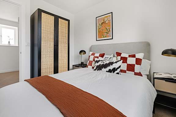 A bed with a white comforter and a brown throw sits in a room with a tall wardrobe and a lamp.