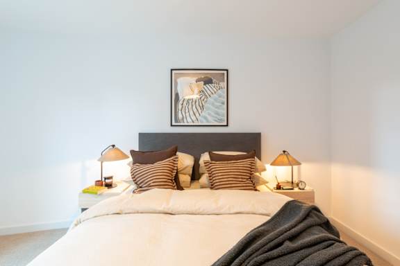 A bed with a white comforter and a striped pillow is in the foreground of a room with a picture frame on the headboard.