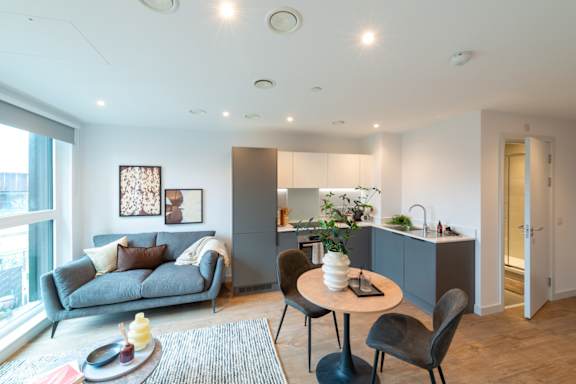 A modern living room with a grey couch, a round table with a vase of flowers, and a kitchen area in the background.