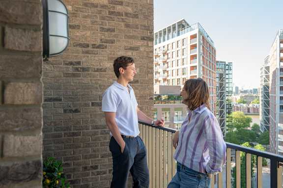 A man and a woman are standing on a balcony with a cityscape in the background.