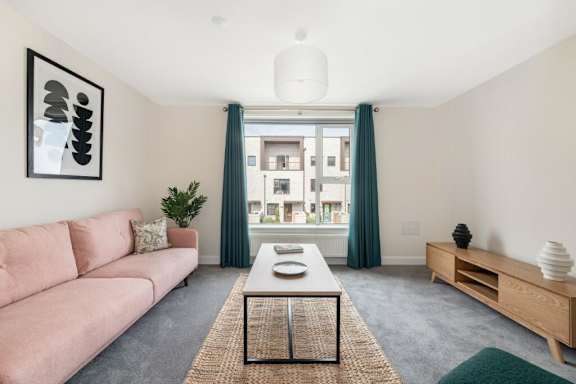 A living room with a pink couch, a coffee table, and a large window with green curtains at Origins Apartments, Cambridge, CB3 0UX