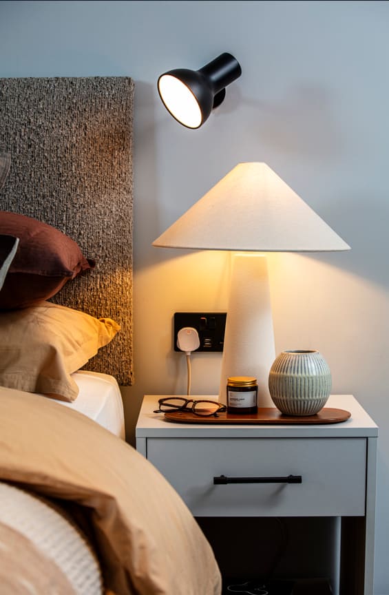 A lamp with a white shade is on a nightstand next to a bed.