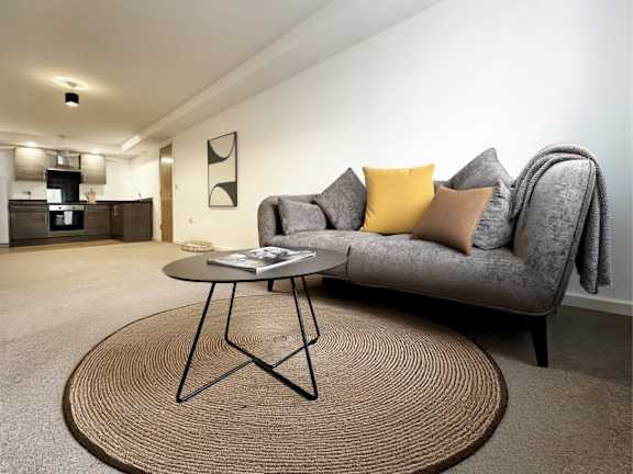 A grey couch with a yellow pillow is in the foreground of a room with a round table and a rug.