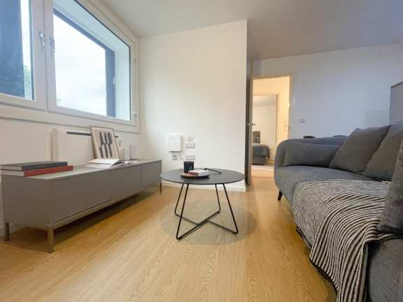 A living room with a grey couch, a table with a cup on it, and a window.