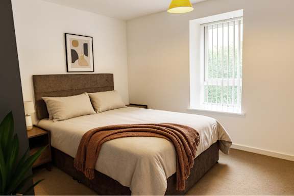 A bedroom with a bed, pillows, a blanket, a nightstand, a plant, and a window with blinds.