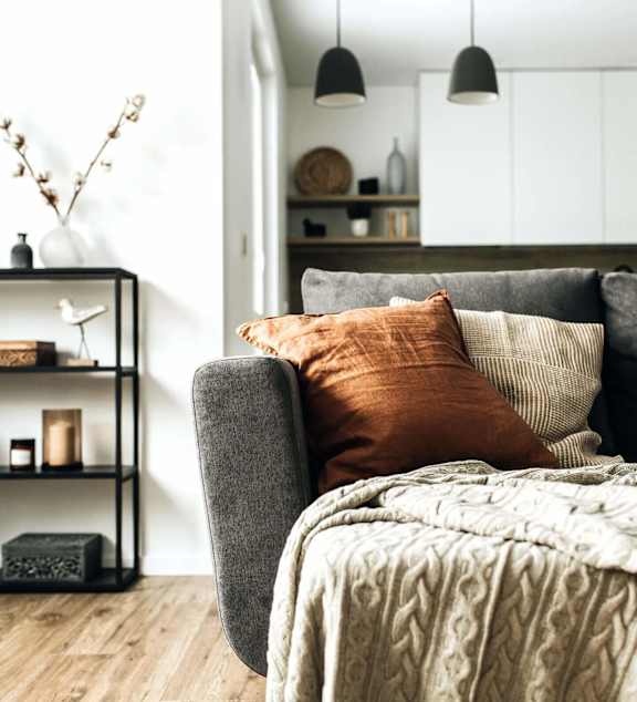 A grey couch with a blanket and pillows in a room with a shelf and decorations.