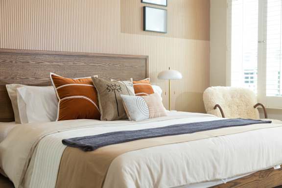 A bed with a white comforter and a blue blanket is in the foreground with a chair and lamp in the background.
