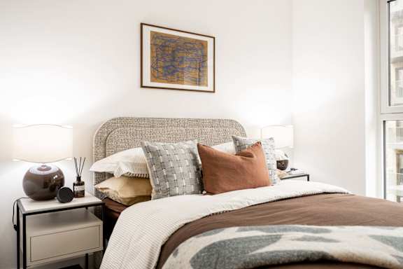 A bedroom with a bed, pillows, a nightstand with a lamp and a vase on it, and a framed picture on the wall.