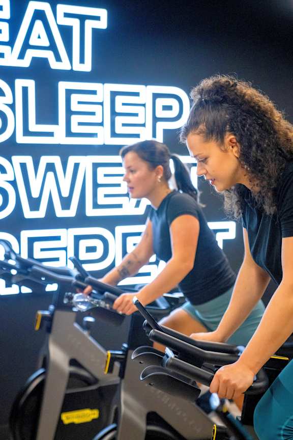 A gym with stationary bikes and a neon sign that says "Eat Sleep Sweat Repeat".