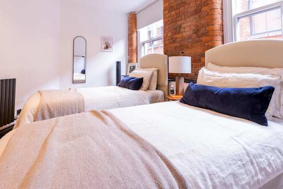 Two beds with white bedspreads and blue pillows in a room with a brick wall.