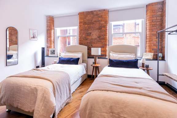 Two twin beds with white bedspreads and beige blankets in a room with brick walls.