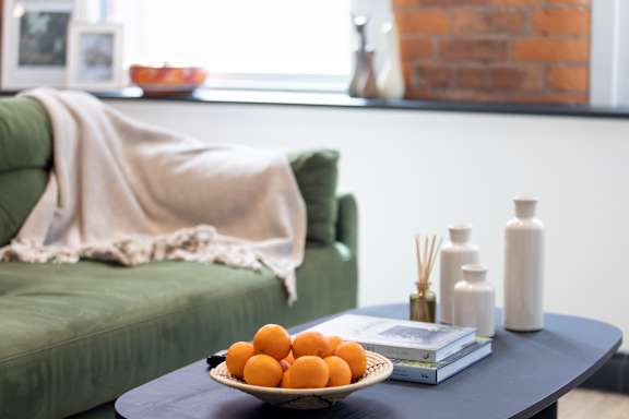 A bowl of oranges sits on a table next to a green couch.