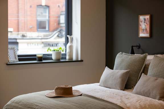 A bedroom with a bed, pillows, a hat, and a window overlooking a city street.