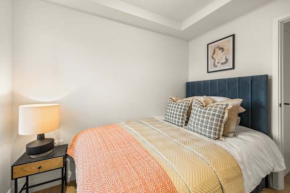 A bed with a white comforter and orange throw is in a room with a lamp and picture on the wall.