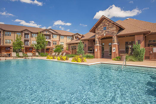 Swimming pool and lounge chairs in front of tan apartment buildings and a stone clubhouse entrance at Cypress at Lewisville