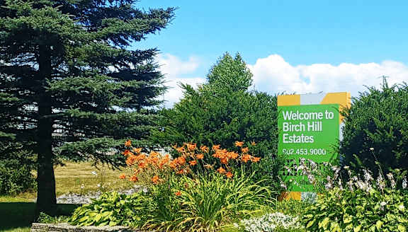 A sign welcoming people to Birch Hill Estates stands in front of a green lawn.
