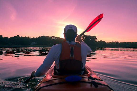 A person wearing a white shirt and a red life vest is kayaking on a calm body of water during sunset.