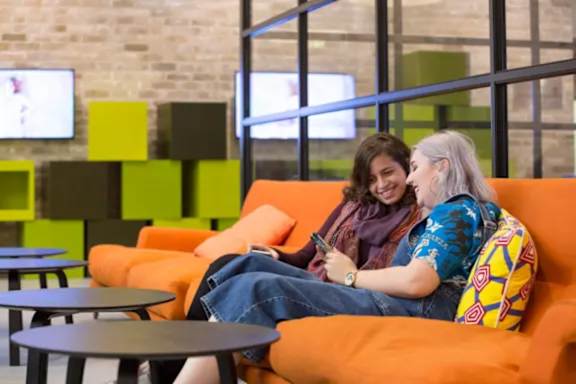 Two people sitting on an orange couch in a room with a brick wall and multiple TVs.
