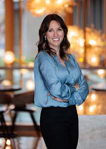 A woman in a blue blouse and black pants stands with her arms crossed.