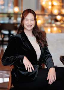 A woman in a black velvet jacket is sitting on a chair.