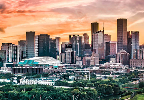 A city skyline at sunset with tall buildings and a stadium in the foreground.
