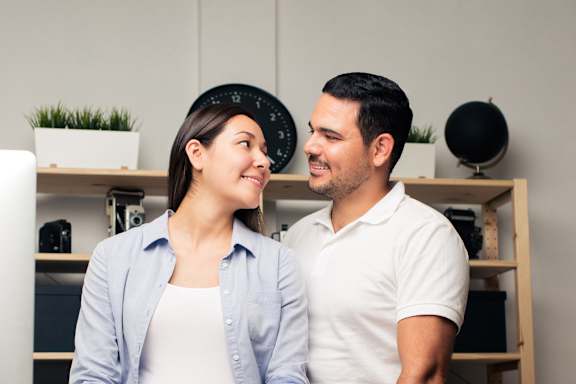 A man and a woman are standing close together in a room with shelves and a clock in the background.