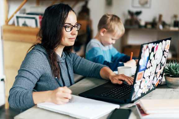 A woman is working on a laptop with a child in the background.