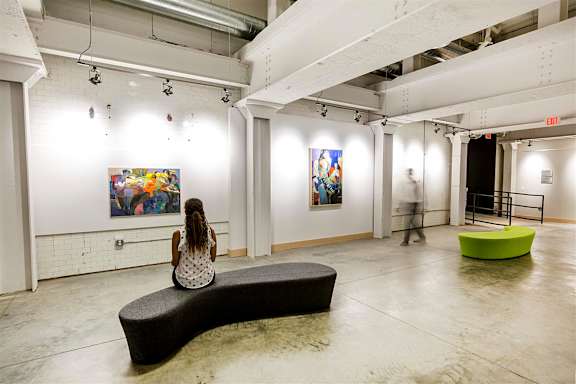 A woman sits on a bench in a gallery looking at paintings.